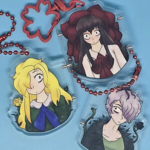 Könnte beinhalten: Drei Schlüsselanhänger im Anime-Stil aus Acryl mit roten Kettenbefestigungen. Jeder Schlüsselanhänger zeigt einen anderen Charakter mit großen Augen und detaillierter Kleidung. Die Charaktere haben blonde, schwarze und lila Haare. Der Hintergrund ist hellblau.