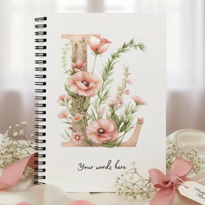 May include: Spiral-bound notebook with a watercolor floral design. The cover features a large letter "I" adorned with pink poppies and greenery. The words "Your words here" are printed at the bottom. A pink ribbon and tag with the words "Happy You Thoughts" are also visible.
