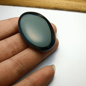 Puede incluir: Una piedra preciosa ovalada y pulida, con un centro verde-azulado oscuro y un borde azul-marrón oscuro. La piedra se sostiene en una mano, mostrando su superficie lisa y reflectante. La forma y el color de la gema sugieren su uso en joyería o como objeto decorativo.