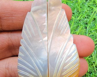 May include: Two symmetrical, leaf-shaped pieces carved from iridescent mother-of-pearl. Each piece features delicate, etched vein patterns across the surface, transitioning from a translucent, pearlescent white at the top to a warm, golden-orange hue at the base.