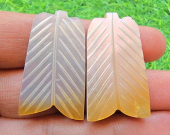 May include: Two translucent, light orange, leaf-shaped stone carvings featuring a central vein and diagonal etched lines. These flat, symmetrical pieces have a smooth, polished surface and a notched base, showcasing a soft, warm, semi-precious appearance.