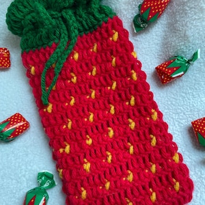 May include: A handmade crochet strawberry-shaped bag with a green drawstring closure. The bag is red with yellow seed details. Several strawberry-wrapped candies are scattered around the bag.