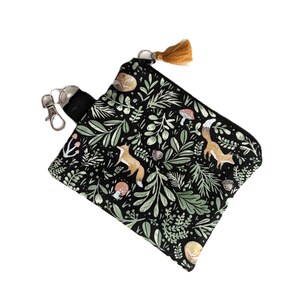 May include: A square zippered pouch featuring a black background with a repeating pattern of orange foxes, green foliage, mushrooms, and acorns. The pouch includes a black fabric strap with a silver metal lobster clasp and a small mustard yellow tassel.