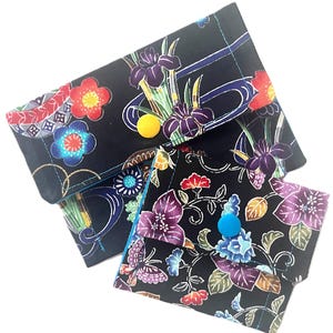May include: Two black fabric pouches with floral patterns. The larger pouch has a yellow button closure, while the smaller one has a blue button. The floral designs include irises, butterflies, and other colorful blooms.