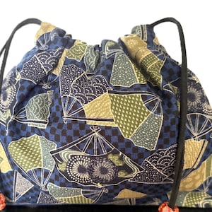 May include: A drawstring bag with a Japanese fan pattern in blue, green, and beige. The bag has black drawstrings and small red and white beads at the ends. The bag is made of fabric.