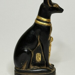 May include: Black and gold Egyptian cat statue with hieroglyphics on the base. The statue is sitting on a square base with hieroglyphics on all sides.