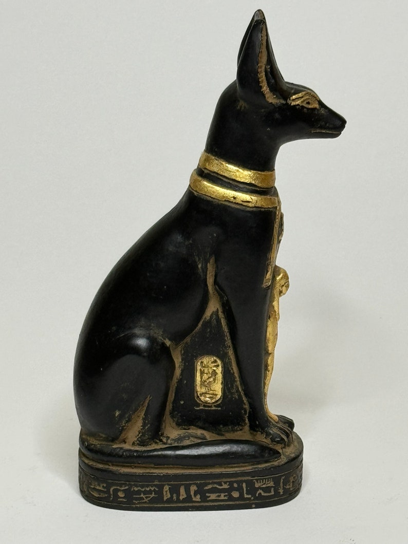 May include: Black and gold Egyptian cat statue with hieroglyphics on the base. The statue is sitting on a square base with hieroglyphics on all sides.