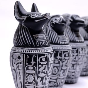 Könnte beinhalten: Reihe von schwarz-weißen ägyptischen Kanopenkrügen. Jeder Krug weist detaillierte Schnitzereien auf, darunter Hieroglyphen und Tierkopf-Figuren. Die Krüge sind in einer Reihe angeordnet, wobei der erste Krug im Fokus steht.