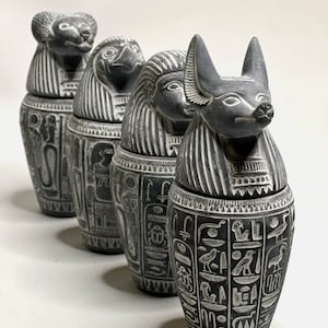 Peut inclure: Quatre jarres canopes en céramique grise avec des hiéroglyphes égyptiens et des couvercles en tête d'animal. Les couvercles sont un chacal, un babouin, une tête humaine et un chien.