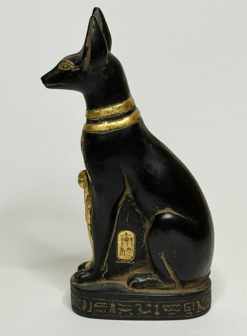 May include: Black and gold Egyptian cat statue with hieroglyphics on the base. The cat is sitting with its head turned to the side.