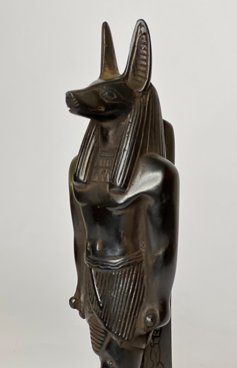 May include: Black statue of the Egyptian god Anubis, depicted as a jackal with a human body. The statue is standing upright with its arms crossed in front of its chest. The statue is made of a dark, smooth material and has a detailed design.