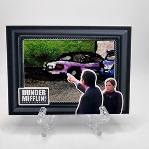 Puede incluir: Una impresión enmarcada de una escena del programa de televisión "The Office" con Michael Scott y Pam Beesly. La impresión está en un marco negro con un passe-partout blanco. La impresión muestra a Michael señalando un coche morado y Pam mirándolo con una expresión confusa. La impresión también incluye el logotipo de Dunder Mifflin.