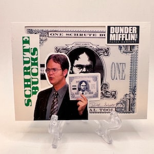 Puede incluir: Una tarjeta blanca con una imagen en blanco y negro de un hombre sosteniendo una versión más pequeña de la tarjeta. La tarjeta tiene el texto "One Schrute Buck" y "Dunder Mifflin Paper Company" impreso en ella. La tarjeta es verde y blanca y tiene un borde verde.