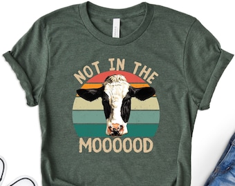 May include: A green t-shirt with a retro-style graphic of a cow's head in front of a sunset. The text "Not in the Mooooood" is printed below the cow.