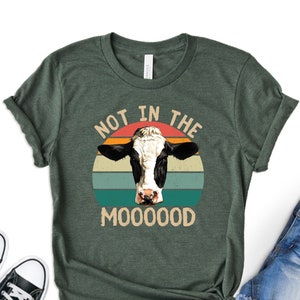 May include: A green t-shirt with a retro-style graphic of a cow's head in front of a sunset. The text "Not in the Mooooood" is printed below the cow.