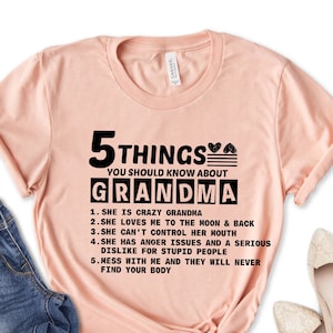 May include: A light pink t-shirt with the text "5 THINGS YOU SHOULD KNOW ABOUT GRANDMA" in black. The text is followed by a list of five humorous statements about grandmothers. The shirt is styled with blue jeans and beige flats.