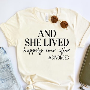 Puede incluir: Una camiseta blanca con el texto "AND SHE LIVED happily ever after #DIVORCED" impreso en negro.