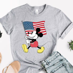 May include: A gray t-shirt with a cartoon image of Mickey Mouse holding an American flag. Mickey Mouse is wearing red shorts and yellow shoes.