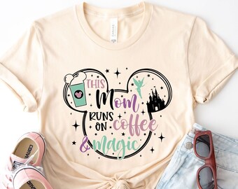 May include: A beige t-shirt with a Mickey Mouse head design. The design includes a coffee cup, a castle, and Tinkerbell. The text on the shirt reads "This Mom Runs on Coffee & Magic".