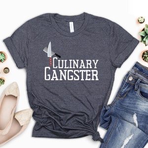May include: Dark gray t-shirt with the text "CULINARY GANGSTER" in white. The shirt features a cleaver and knife graphic. A pair of blue jeans and beige flats are next to the shirt.