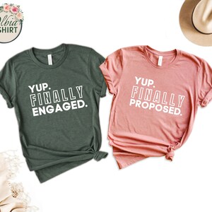 May include: Two t-shirts, one dark green and one dusty rose, with white text. The dark green shirt says "Yup. Finally Engaged." The dusty rose shirt says "Yup. Finally Proposed."