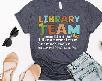 May include: A dark blue t-shirt with the text "LIBRARY TEAM" in colorful letters. The text "noun/librerë tëm/ 1.like a normal team, but much cooler. (see also: best friends, exceptional)" is printed below the main text.
