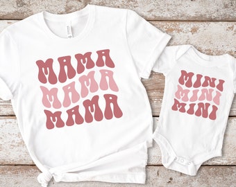 May include: White t-shirt with the text "MAMA MAMA MAMA" in pink and red, and a white bodysuit with the text "MINI MINI MINI" in pink and red.