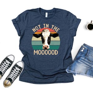 May include: A navy blue t-shirt with a retro-style graphic of a cow's head in front of a sunset. The text "Not in the Mooooood" is printed below the graphic.