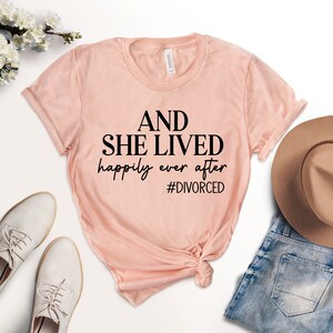 Puede incluir: Una camiseta rosa con el texto "AND SHE LIVED happily ever after #DIVORCED" impreso en negro.
