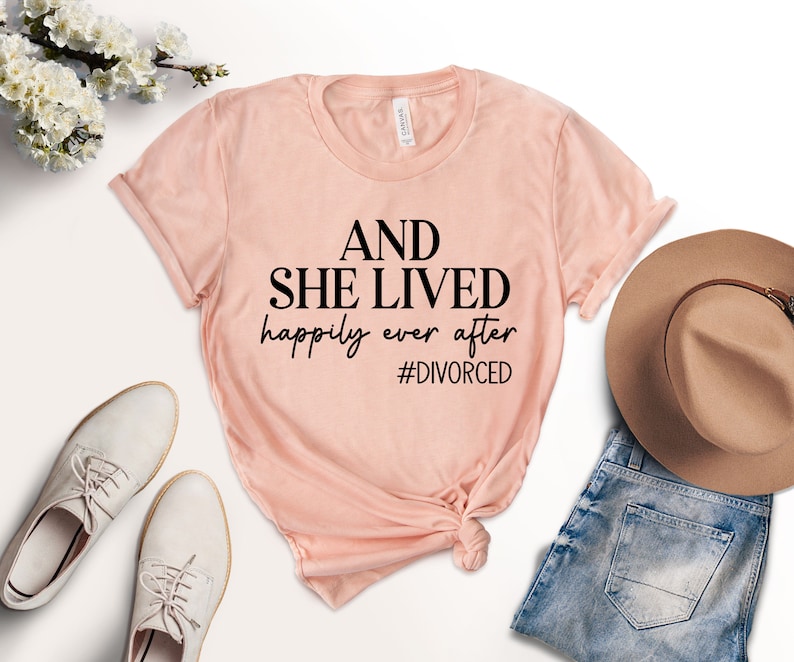 Puede incluir: Una camiseta rosa con el texto "AND SHE LIVED happily ever after #DIVORCED" impreso en negro.