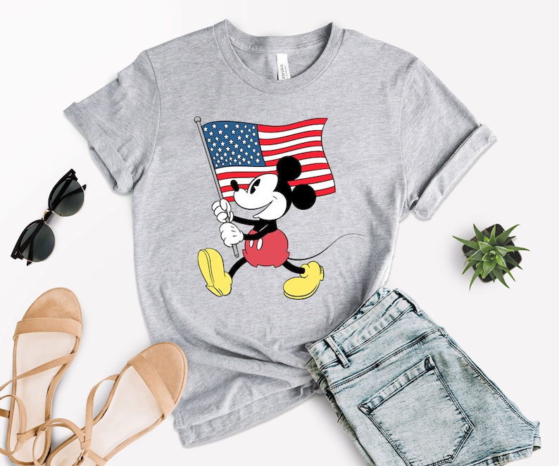 May include: A gray t-shirt with a cartoon image of Mickey Mouse holding an American flag. Mickey Mouse is wearing red shorts and yellow shoes.