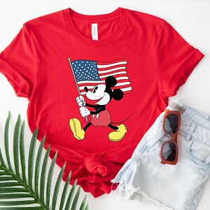 May include: Red t-shirt with a graphic of Mickey Mouse holding an American flag. The shirt is made of a soft, comfortable material.