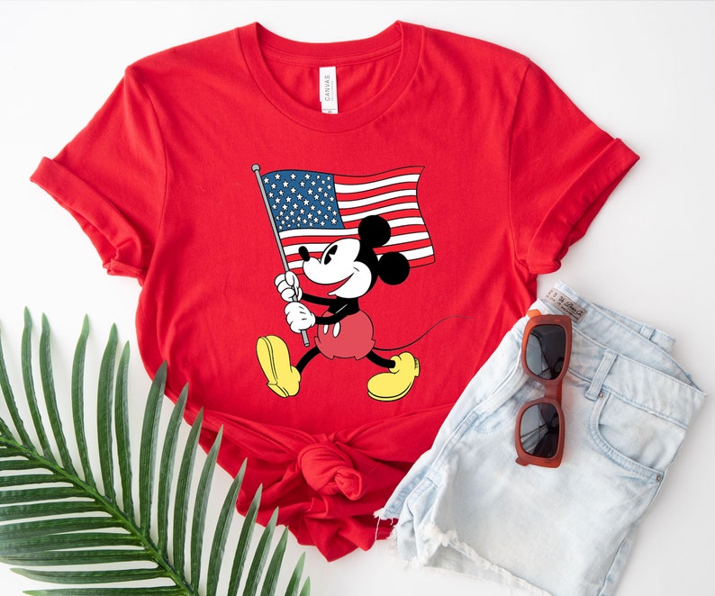 May include: Red t-shirt with a graphic of Mickey Mouse holding an American flag. The shirt is made of a soft, comfortable material.