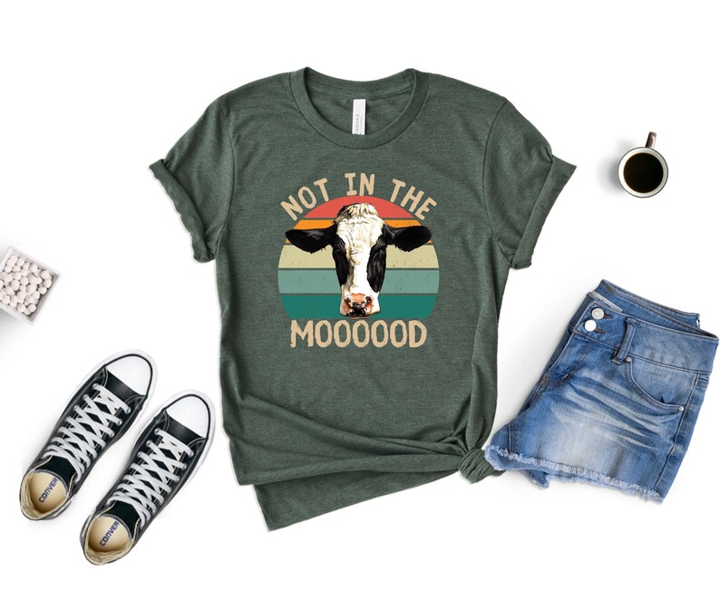 May include: A green t-shirt with a retro-style graphic of a cow's head in front of a sunset. The text "Not in the Mooooood" is printed below the cow.
