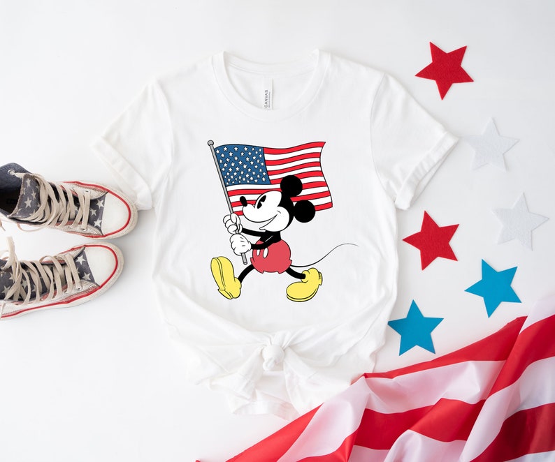 May include: White t-shirt with a cartoon image of Mickey Mouse holding an American flag. The shirt is tied in a knot at the bottom. There are red, white, and blue stars scattered around the shirt.