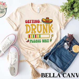 May include: A beige t-shirt with the text "Getting Drunk Please Wait" in a colorful font. The text is stylized to look like a progress bar. A sombrero is above the text. The shirt is paired with blue denim shorts.