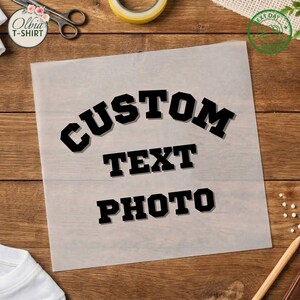 May include: A square, semi-transparent sheet featuring the black, bold, block-lettered text CUSTOM TEXT PHOTO arranged in three lines. The sheet is positioned on a wooden surface alongside a pair of scissors and a yellow tape roll.