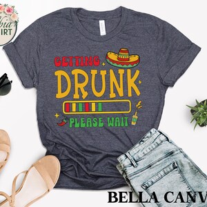 May include: A dark blue t-shirt with the text "Getting Drunk" and a progress bar that says "Please Wait". The progress bar is filled with green, red, and yellow stripes. There is a sombrero above the text and a chili pepper and a margarita glass below the text.