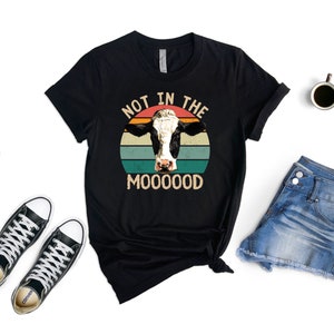 May include: Black t-shirt with a vintage-style graphic of a cow's head in front of a sunset. The text "Not in the Mooood" is printed below the cow.