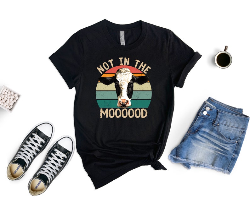 May include: Black t-shirt with a vintage-style graphic of a cow's head in front of a sunset. The text "Not in the Mooood" is printed below the cow.