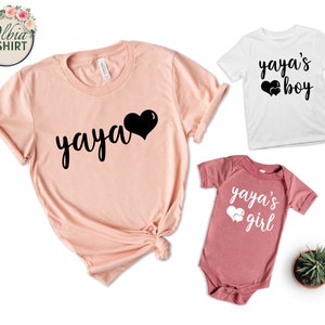 K&ouml;nnte beinhalten: Ein Set aus drei T-Shirts mit dem Text "yaya" und Herzen. Das gr&ouml;&szlig;te Shirt ist pfirsichfarben und hat den Text "yaya" in Schwarz mit einem schwarzen Herzen. Die kleineren Shirts sind wei&szlig; und rosa und haben den Text "yaya's boy" und "yaya's girl" in Schwarz mit wei&szlig;en Herzen.