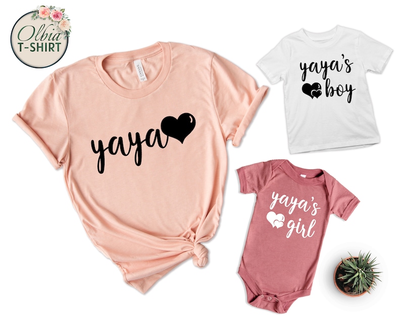 K&ouml;nnte beinhalten: Ein Set aus drei T-Shirts mit dem Text "yaya" und Herzen. Das gr&ouml;&szlig;te Shirt ist pfirsichfarben und hat den Text "yaya" in Schwarz mit einem schwarzen Herzen. Die kleineren Shirts sind wei&szlig; und rosa und haben den Text "yaya's boy" und "yaya's girl" in Schwarz mit wei&szlig;en Herzen.
