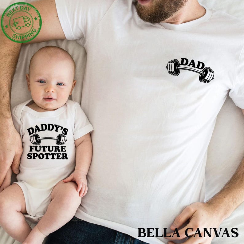 Pu&ograve; includere: Un body bianco con il testo "Daddy's Future Spotter" e una grafica di bilanciere nero. Una maglietta bianca con il testo "DAD" e una grafica di bilanciere nero. Il body &egrave; indossato da un bambino e la maglietta &egrave; indossata da un adulto.