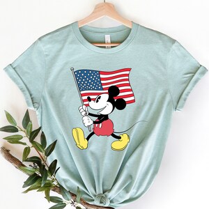 May include: A light blue t-shirt with a graphic of Mickey Mouse holding an American flag. Mickey Mouse is wearing red shorts and yellow shoes.