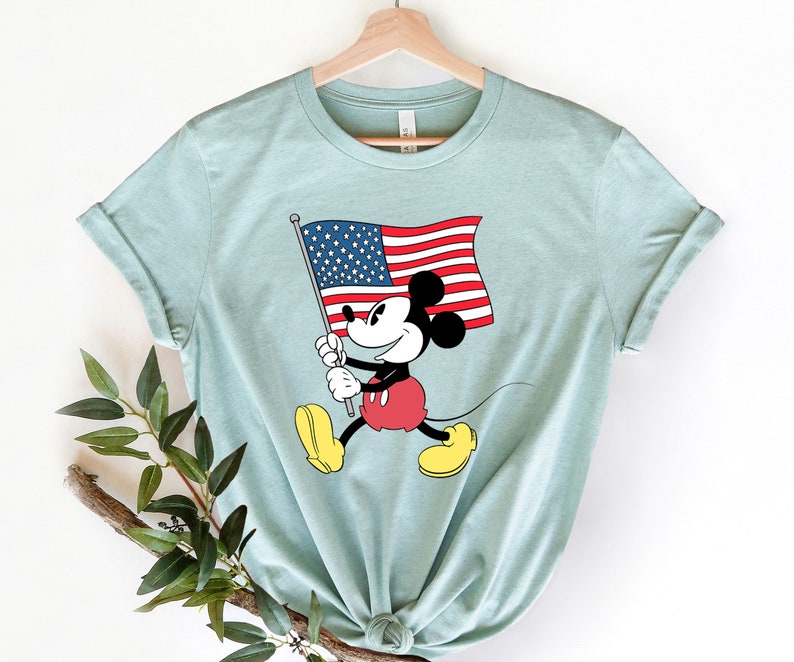 May include: A light blue t-shirt with a graphic of Mickey Mouse holding an American flag. Mickey Mouse is wearing red shorts and yellow shoes.