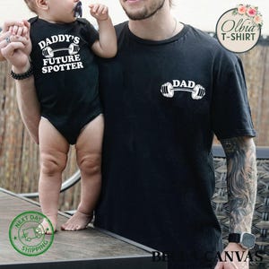 Pu&ograve; includere: Una maglietta nera con la parola "DAD" stampata su di essa, con un bilanciere sotto. Un bambino indossa un body nero con il testo "Daddy's Future Spotter" e un bilanciere sotto.