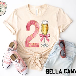 May include: A beige t-shirt with a pink and gold number 2 and a glass of champagne with a pink ribbon. The shirt is on a white background with a pair of pink and white sneakers and a pair of brown sunglasses.