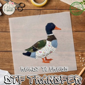 May include: A DTF transfer sheet featuring a mallard duck graphic composed of various fabric patterns including camouflage, floral lace, and solid blue patches. The duck has a dark head, orange beak, and orange feet. The text READY TO PRESS is below.
