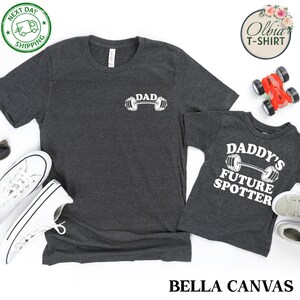 Pu&ograve; includere: Due magliette grigie con testo bianco e un grafico di un bilanciere. La maglietta pi&ugrave; grande dice "DAD" e quella pi&ugrave; piccola dice "DADDY'S FUTURE SPOTTER".