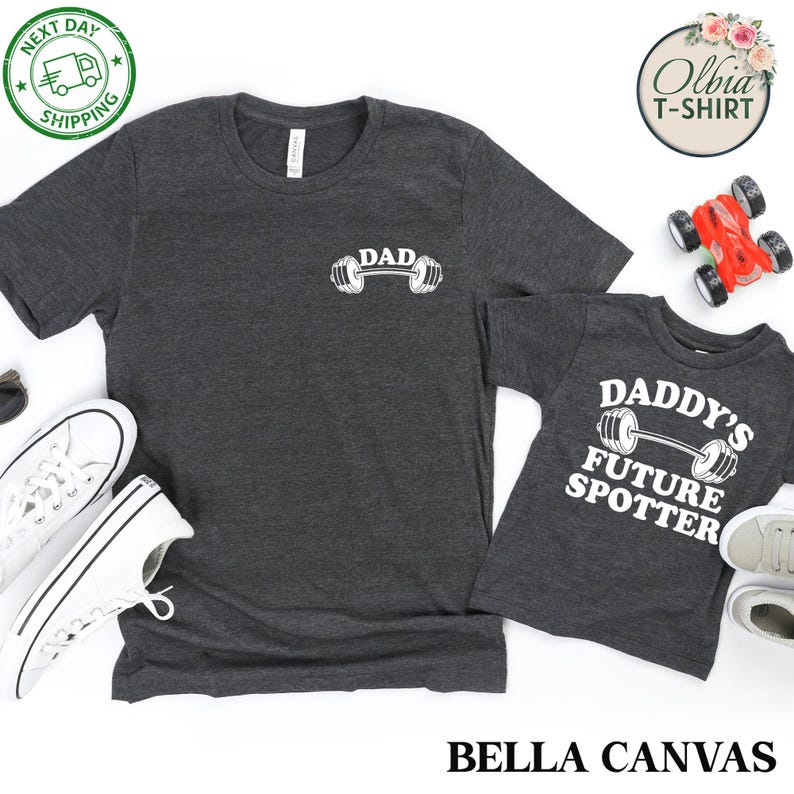 Pu&ograve; includere: Due magliette grigie con testo bianco e un grafico di un bilanciere. La maglietta pi&ugrave; grande dice "DAD" e quella pi&ugrave; piccola dice "DADDY'S FUTURE SPOTTER".